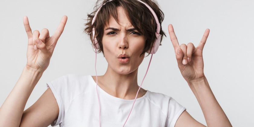 Image of happy woman in basic t-shirt showing rock sign while listening to music with headphones isolated over white background